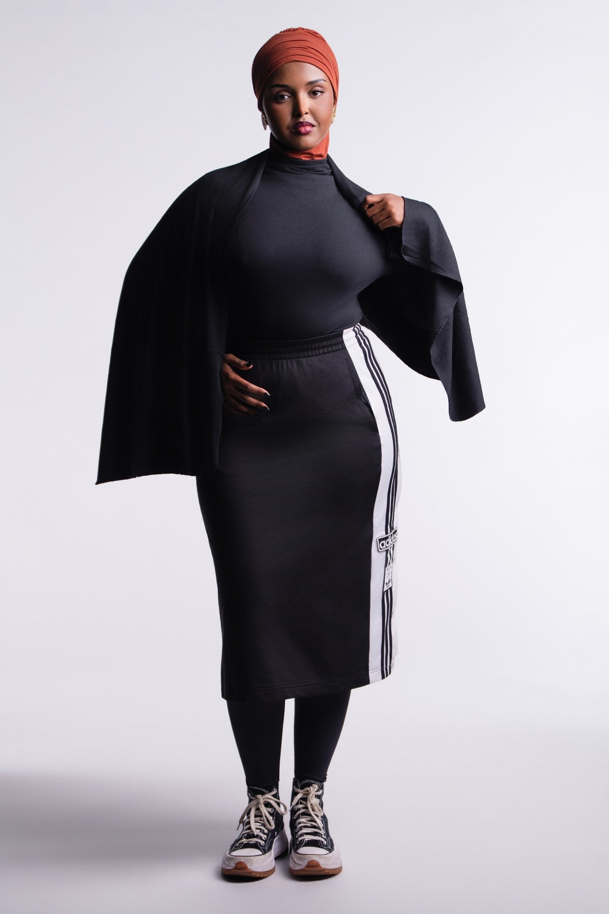Front view of the Black Convertible Veil Bodysuit with the overlay draped over the shoulders as a shawl, styled with a black track skirt and sneakers.
