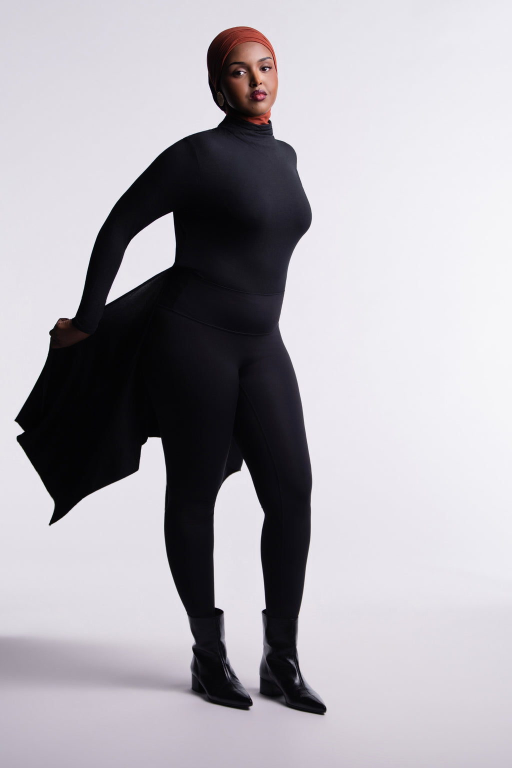 Front view of the Black Convertible Veil Bodysuit with the overlay worn open and swept to one side, showing the fitted mock-neck bodysuit, styled with black leggings and ankle boots.