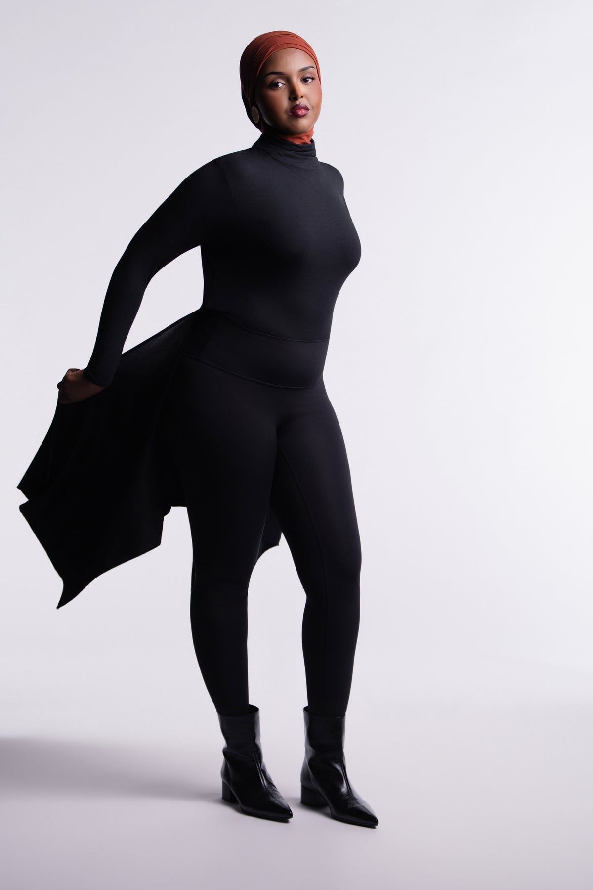 Front view of the Black Convertible Veil Bodysuit with the overlay worn open and swept to one side, showing the fitted mock-neck bodysuit, styled with black leggings and ankle boots.
