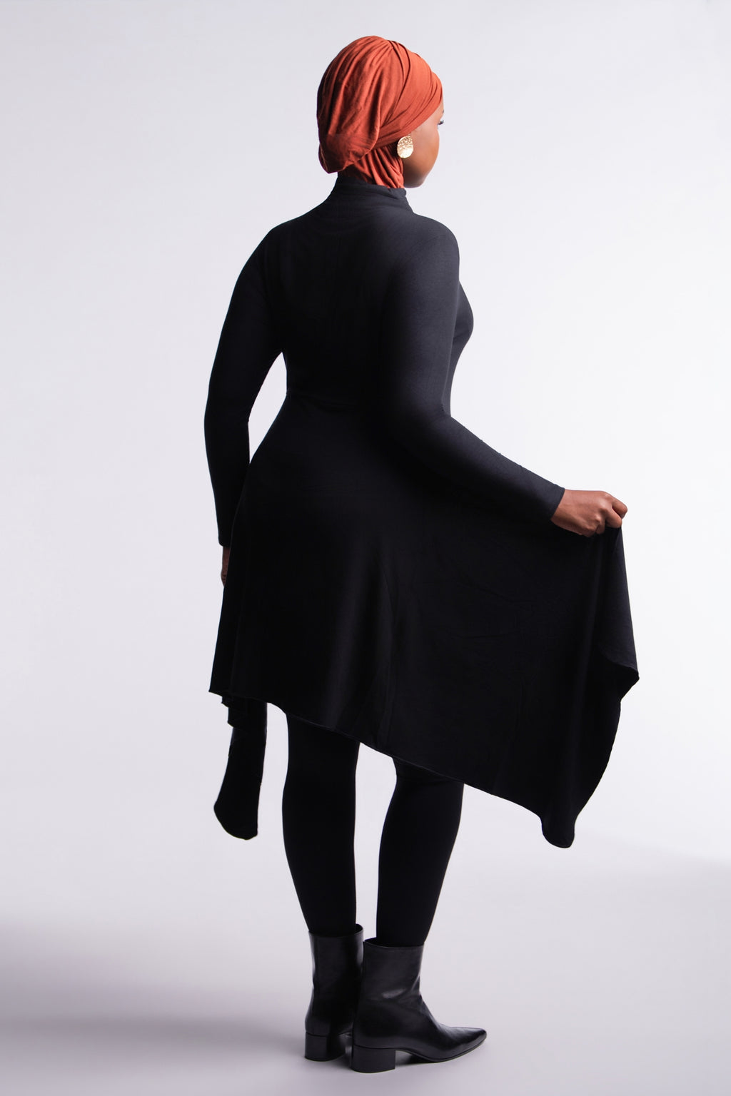 Back view of the Black Convertible Veil Bodysuit with the overlay worn closed as a tunic, showing the flowing asymmetric hem, styled with black leggings and ankle boots.