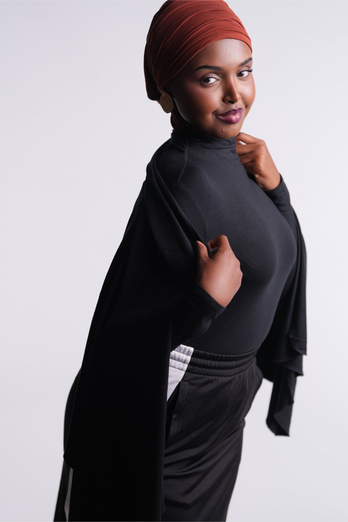 DuoWear 2-in-1 Hijab worn turban style in a rich rust brown, with the black Convertible Veil Bodysuit draped as a shawl and black track pants