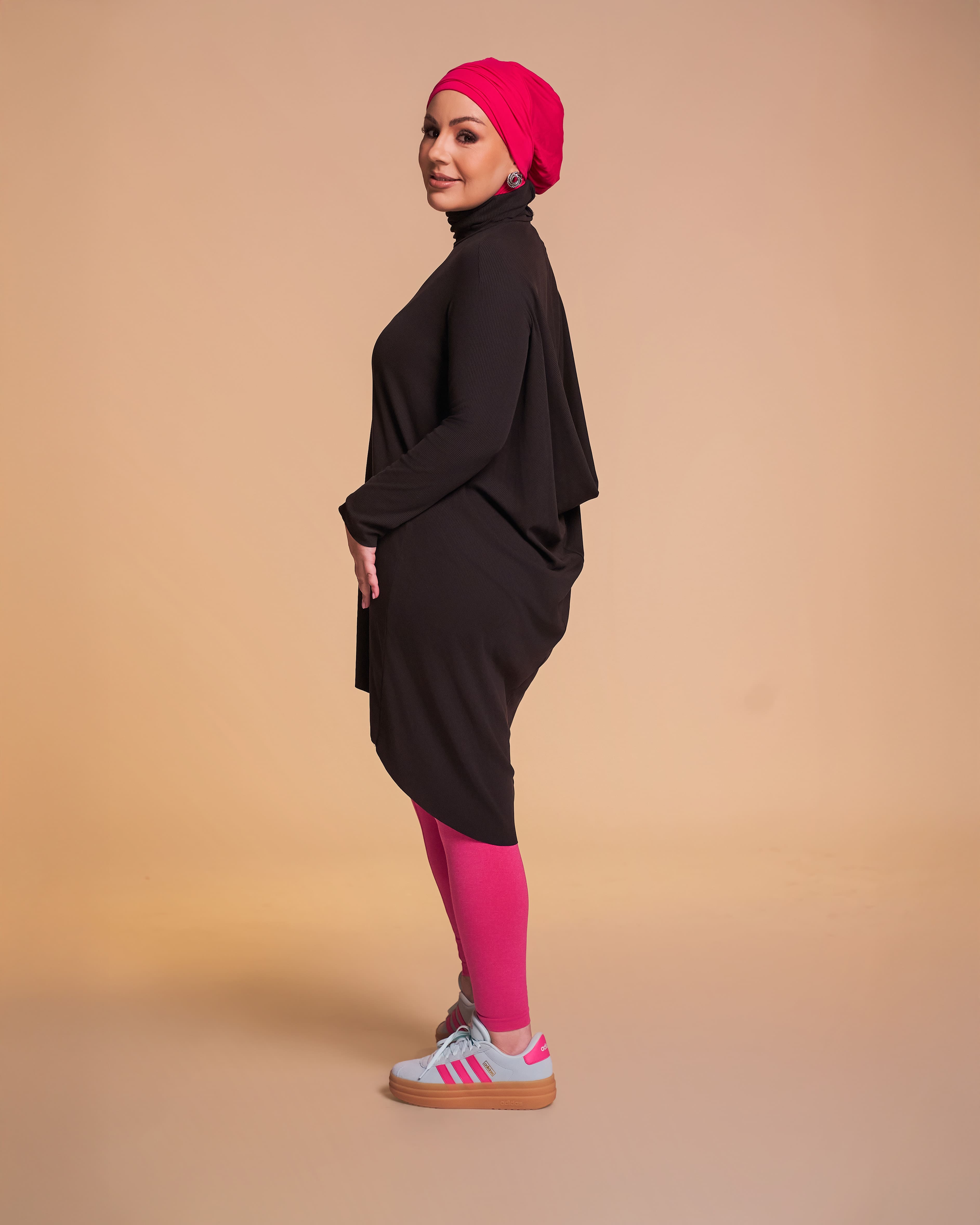 Studio photo of a woman wearing the DuoWear Hijab in hot pink and the Flow Tunic in black styled with workout leggings and sneakers.