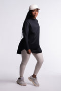Black | Limitless Top side view, mock neck and tunic length with curved hem, worn with a cream cap, beige leggings and chunky sneakers