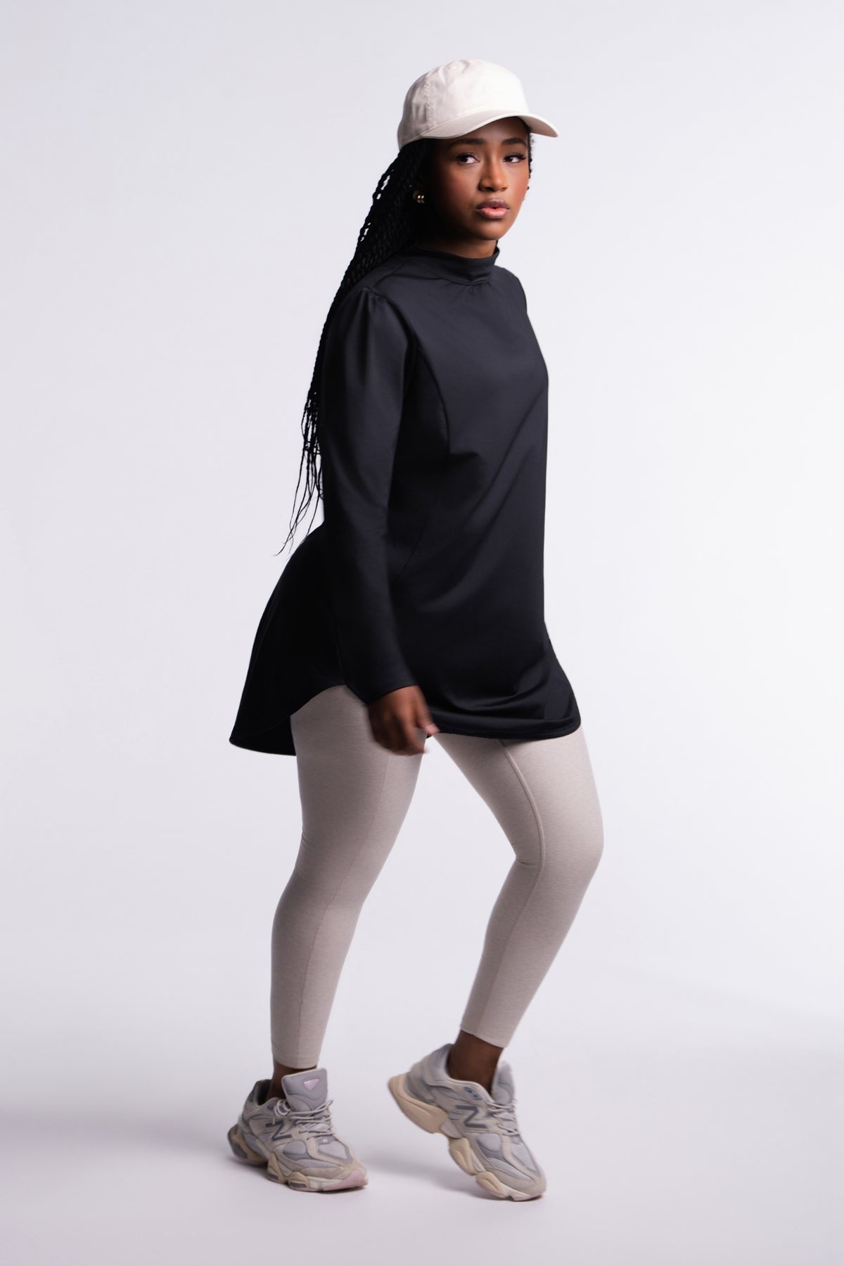 Black | Limitless Top side view, mock neck and tunic length with curved hem, worn with a cream cap, beige leggings and chunky sneakers