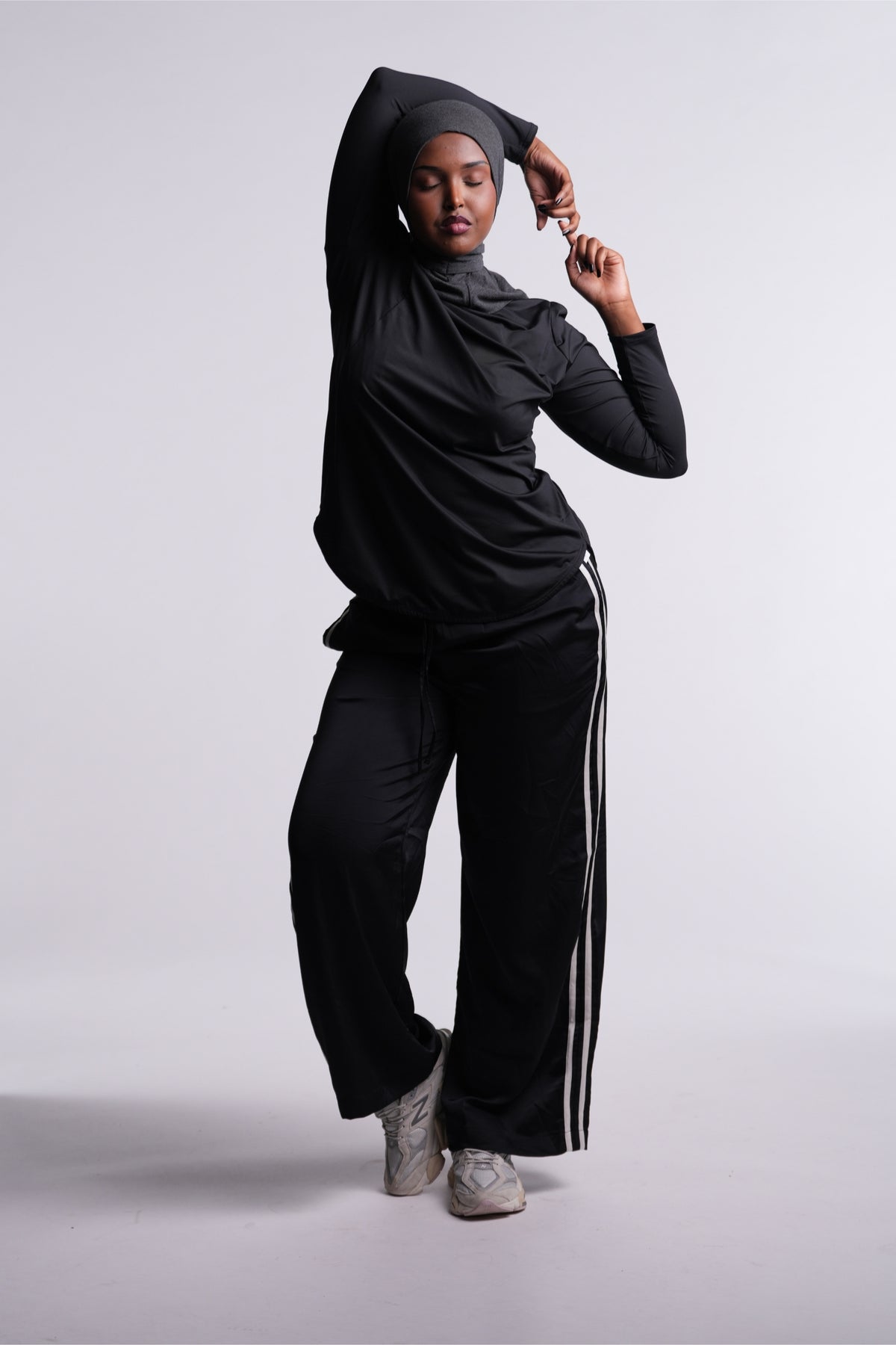 Black | Limitless Top with one arm raised overhead to show stretch, worn with a gray hijab, black track pants and sneakers