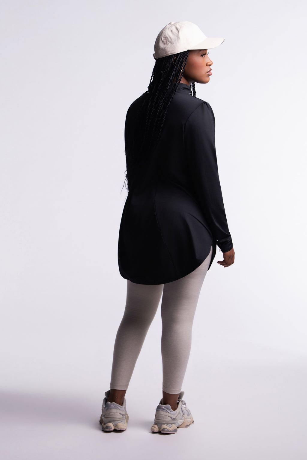 Limitless Top back view, mock neck and tunic length with curved hem, worn with a cream cap, beige leggings and chunky sneakers