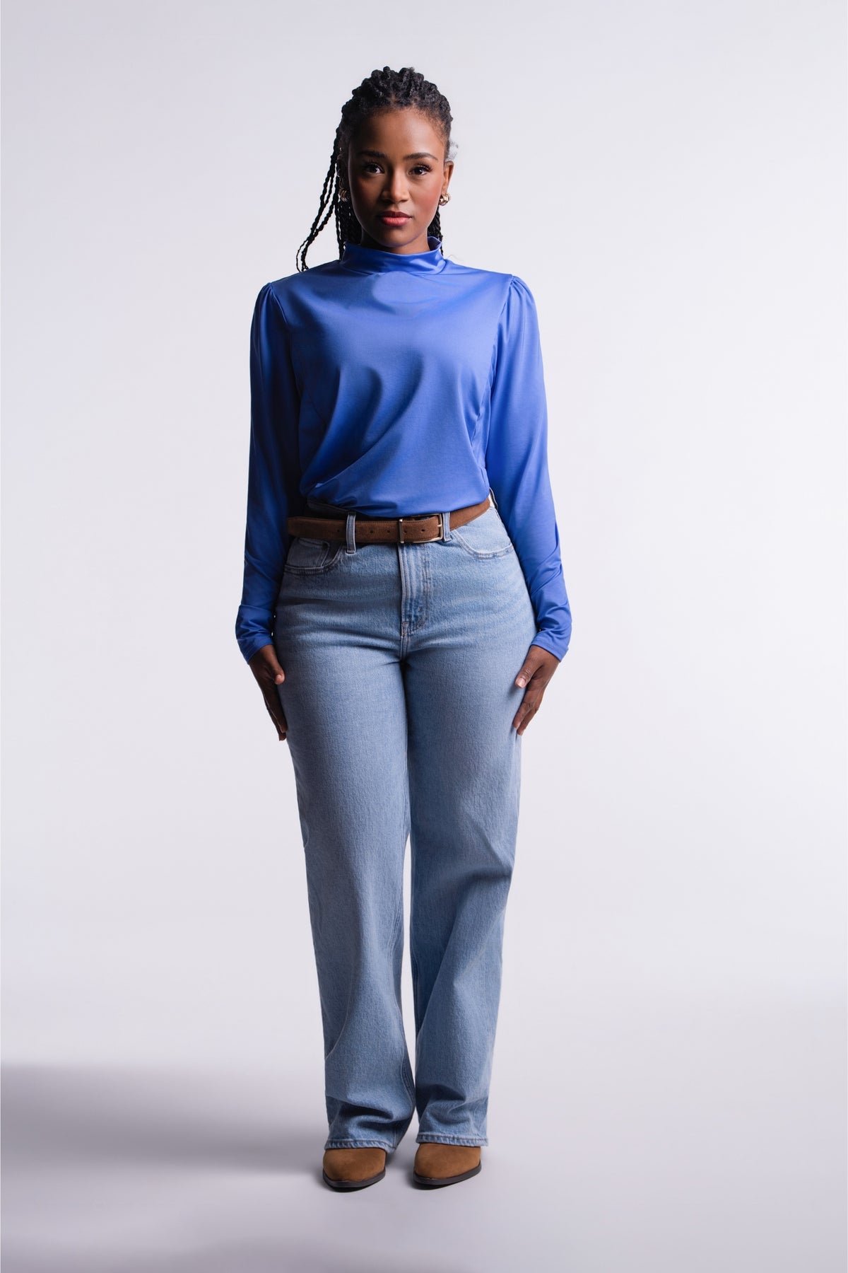 Periwinkle | Limitless Top in periwinkle blue, front view with mock neck and ruched shoulders, tucked into light-wash jeans with a brown belt