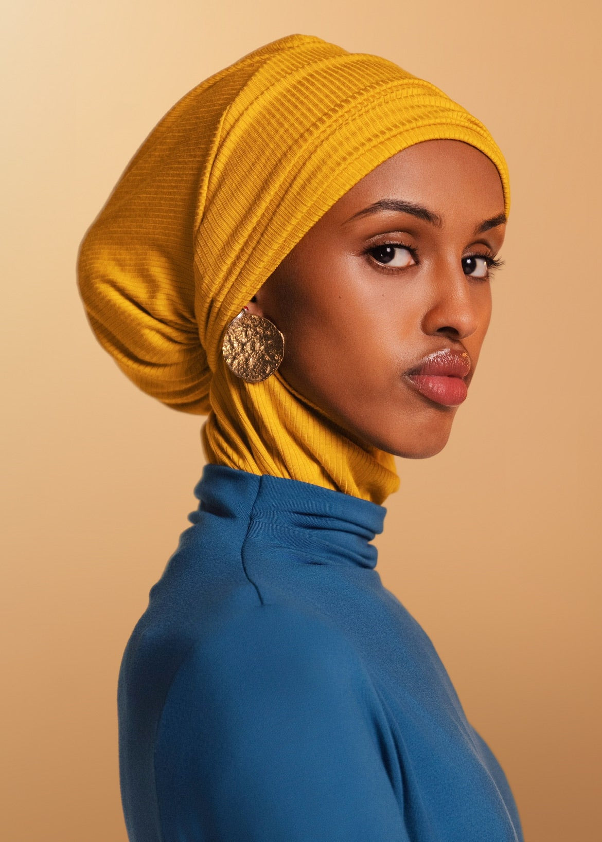 Woman wearing a yellow headscarf and blue top against a beige background