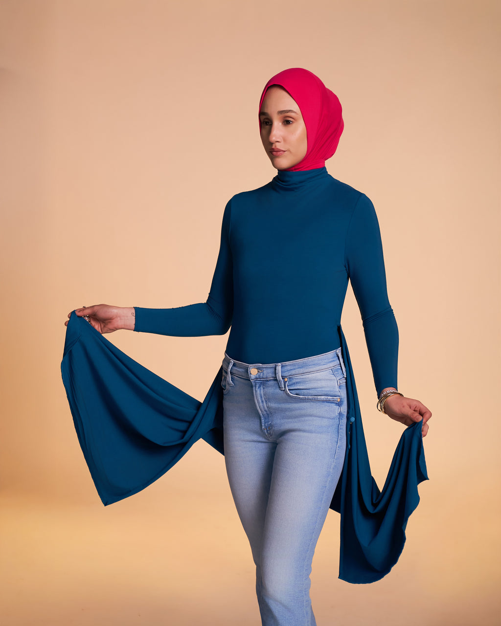 A Woman wearing the Solo Snap Hijab in hot pink and the Veil Bodysuit in teal holding the tunic overlay with her hands.